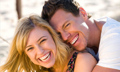 Picture of a smiling couple, happy with the prices they received for dental work at the Costa Rica Dental Centre in San Jose, Costa Rica.  The woman has sandy blonde hair, the man has short dark hair and both are hugging and showing their perfect teeth while smiling directly at the camera.